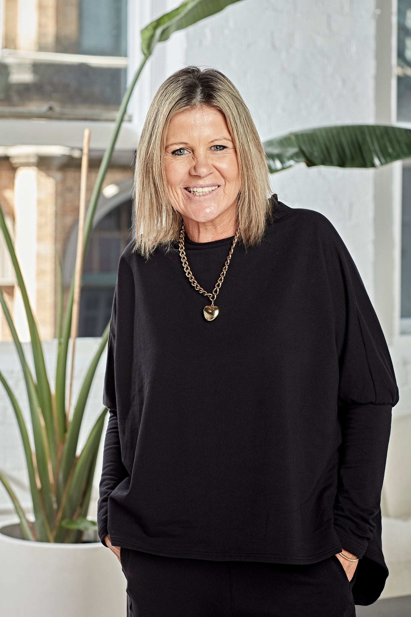 A smiling blonde woman wearing a black long-sleeve top and black pants, accessorized with a gold chain necklace with a heart pendant, standing in front of a white wall with a potted plant.