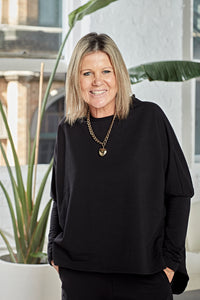 A smiling blonde woman wearing a black long-sleeve top and black pants, accessorized with a gold chain necklace with a heart pendant, standing in front of a white wall with a potted plant.
